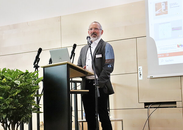 Ein Mann im Anzug mit Brille und Bart steht an einem Rednerpult und hält einen Vortrag.