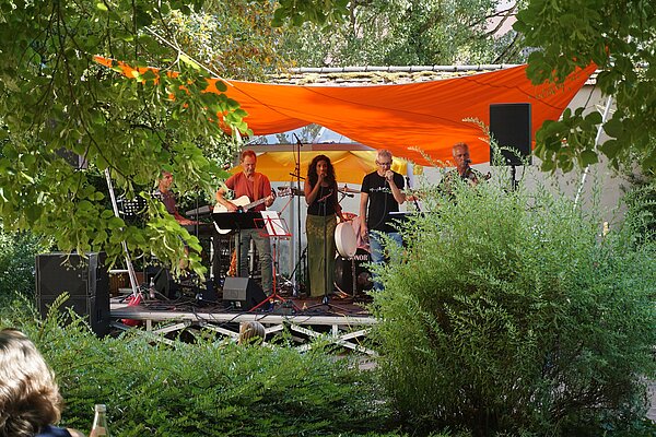 auf einer Bühne mit einem orangefarbenen Segel darüber sind vier Männer mit verschiedenen Musikinstrumenten. Eine Frau hält ein Mikrofon und singt. Im Vordergrund rund um die Bühne befinden sich Bäume und Büsche. 