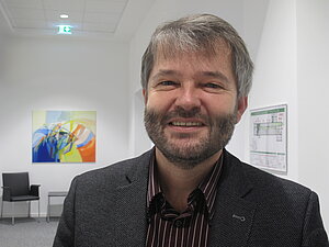 Portraitbild Dr. Matthias Köhler. Ein Mann mittleren Alters mit Vollbart und dunkelrotem Hemd und braunem Sakko, lächelt freundlich mit geöffnetem Mund.