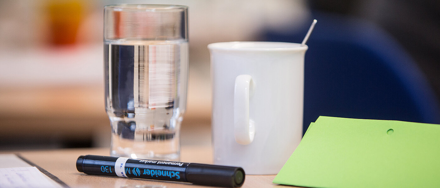 Auf einem Tisch in Nahaufnahme liegen ein schwarzer Textmarker und ein hellgrünes zusammengefaltetes Blatt Papier, außerdem steht auf dem Tisch ein gut gefülltes Glas Wasser sowie eine weiße Kaffeetasse.
