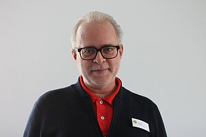 Portraitbild Dr. Thomas Käser, ein Mann fortgeschrittenes Alters mit schwarzer Brille, grauen Haaren und einem grauen Dreitagebart. Er Trägt ein rotes Polo und eine dunkelblaue Strickweste darüber. An der Stickweste auf Brusthöhe hat er ein ZfP-Schildchen angebracht.