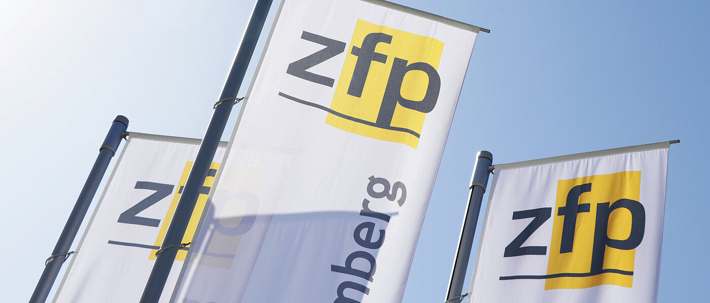 Drei weiße Fahnen mit jeweils dem gelb-schwarzen Logo mit Schriftzug des ZfP Südwürttemberg, im Hintergrund blauer Himmel und Sonnenschein.