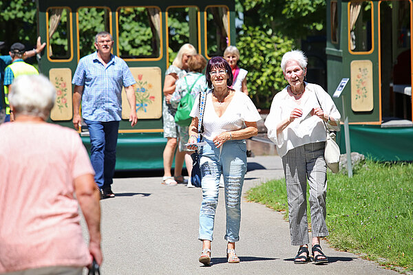 Auf einem geteerten Weg auf dem ZfP-Gelände in Weissenau sind Seniorinnen und Senioren unterwegs vom und zum ZfP-Zügle, das im Hintergrund für die nächste Rundfahrt bereitsteht.