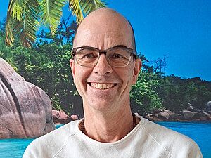 Ein Mann mit Brille und weißem Pullover steht lächelnd vor einer Südsee-Fototapete.