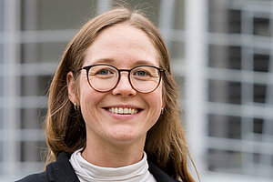 Porträtfoto einer jungen Frau. Sie hat lange, glatte braune Haare mit Seitenscheitel und trägt eine Brille.