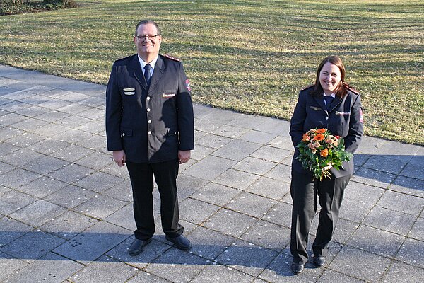 links steht ein Mann mit Brille in Feuerwehruniform, rechts daneben steht eine junge Frau mit braunen offenen Haaren. Sie trägt ebenfalls Feuerwehruniform und hält einen Blumenstrauß in den Händen. Beide lächeln in die Kamera. 