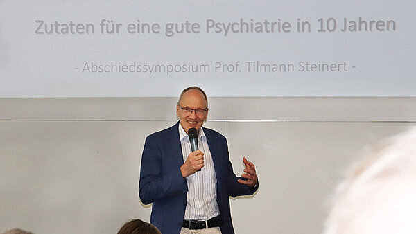 Ein Mann mit Mikrofon in der Hand steht vor einem Menschen in einem Hörsaal.