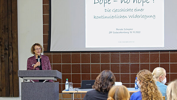 Eine Frau am Rednerpult stehend mit Mikrofon in der Hand hält vor mehreren Zuhörinnen und Zuhörern einen Vortrag, rechts hinter ihr auf einer Leinwand ist eine Powerpoint-Präsentation zu sehen.
