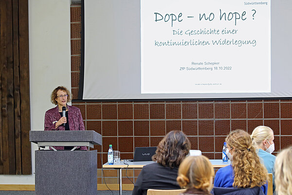 Eine Frau am Rednerpult stehend mit Mikrofon in der Hand hält vor mehreren Zuhörinnen und Zuhörern einen Vortrag, rechts hinter ihr auf einer Leinwand ist eine Powerpoint-Präsentation zu sehen.