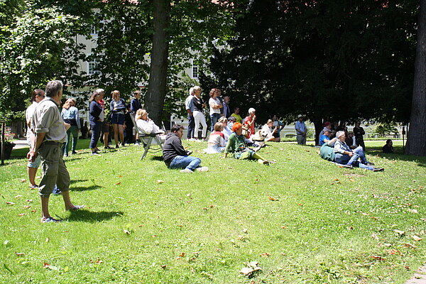 Auf einer Wiese sitzen, liegen und stehen einige Personen, sie schauen alle in die selbe Richtung.