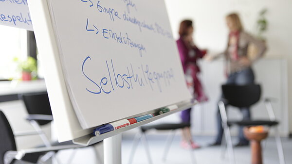 Im Vordergrund ist ein beschriftetes Flipchart zu erkennen, im Hintergrund unterhalten sich zwei Frauen.