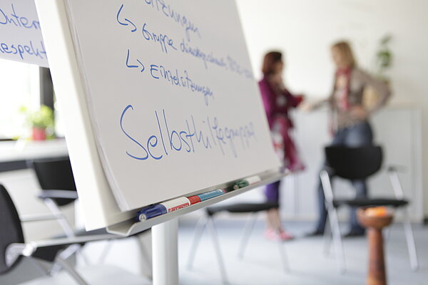 Im Vordergrund ist ein beschriftetes Flipchart zu erkennen, im Hintergrund unterhalten sich zwei Frauen. 