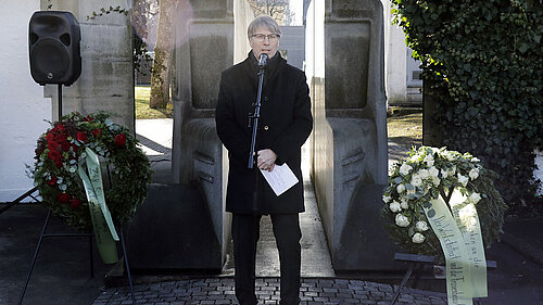 Ein dunkelblonder Mann mittleren Alters im schwarzen Mantel und mit Brille steht an einem Mikrofon und spricht. Im Hintergrund ist ein steinernes Denkmal in Form eines Omnibusses sowie links und rechts zwei Gedenkkränze zu sehen.