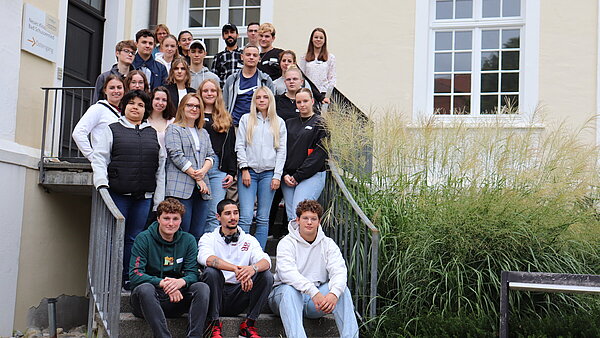 Auf einer Treppe vor einem Gebäude haben sich mehrere junge Menschen für ein Gruppenfoto aufgestellt.
