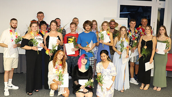 Eine Gruppe junger Menschen steht vor einer weißer Wand. Alle haben eine Rose und eine Urkunde in der Hand.