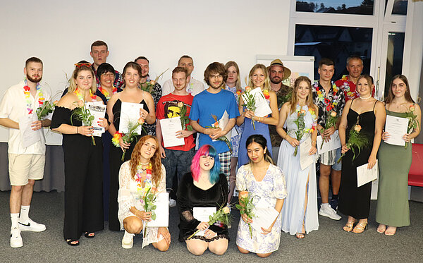 Eine Gruppe junger Menschen steht vor einer weißer Wand. Alle haben eine Rose und eine Urkunde in der Hand.