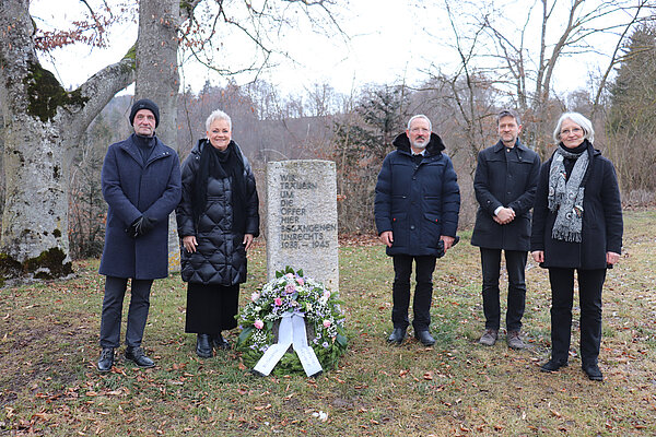 An einem Gedenkstein auf einer Wiese stehen rechts und links verschiedene Personen. An dem Stein liegt ein Gedenkkranz. Im Hintergrund sind Bäume.