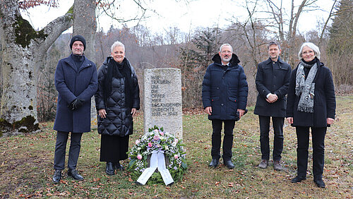 An einem Gedenkstein auf einer Wiese stehen rechts und links verschiedene Personen. An dem Stein liegt ein Gedenkkranz. Im Hintergrund sind Bäume.