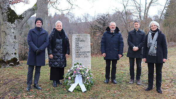 An einem Gedenkstein auf einer Wiese stehen rechts und links verschiedene Personen. An dem Stein liegt ein Gedenkkranz. Im Hintergrund sind Bäume.