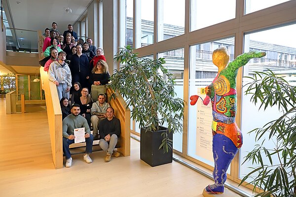 Auf einer langen Treppe im Inneren eines hellen Klinikgebäudes sitzen und stehen etwa 25 Frauen und Männer in Alltagskleidung und lächeln in Richtung Kamera.