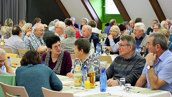 In einem Festsaal sitzen an mehreren festlich gedeckten Tischreihen ältere Damen und Herren beisammen und unterhalten sich miteinander.
