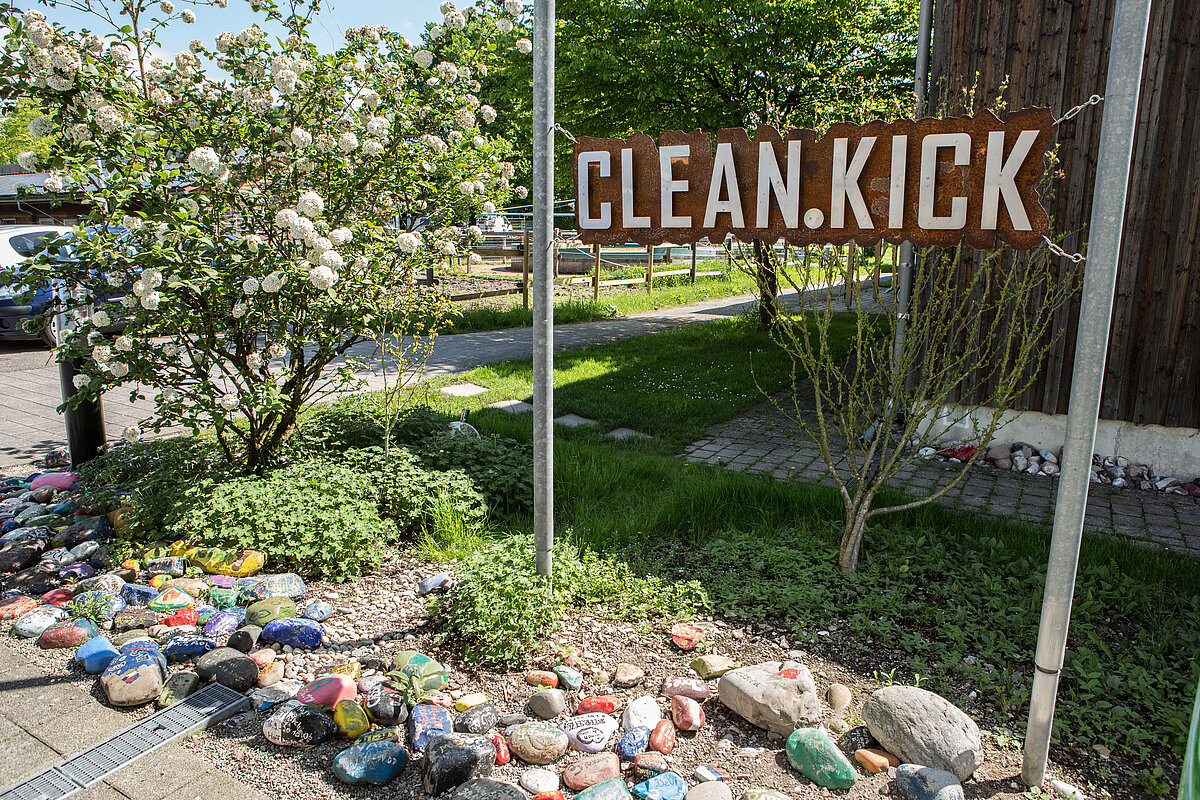 Zugang im Außenbereich zur der Station clean.kick mit Schild, buntbemalten Steinen am Boden und Gras und Bäumen im Hintergrund.