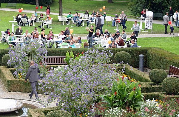 blühender Garten, im Hintergrund sitzen fröhliche Personen an Tischen mit Luftballons