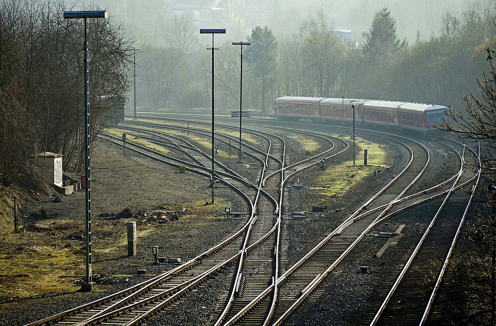 Im Vordergrund ist ein verzweigtes Schienennetz zu sehen, im Hintergrund erkennt man einen roten Zug. 