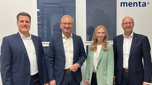 Drei Männer in Anzügen und eine blonde Frau mit hellgrünem Jacket stehen nebeneinander. 