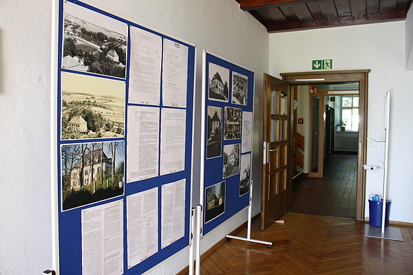 An einer Wand stehen zwei große blaue Stellwände mit weißen Ständern. Auf den Stellwänden sind mehrere historische Gebäudefotos und Textseiten angebracht.