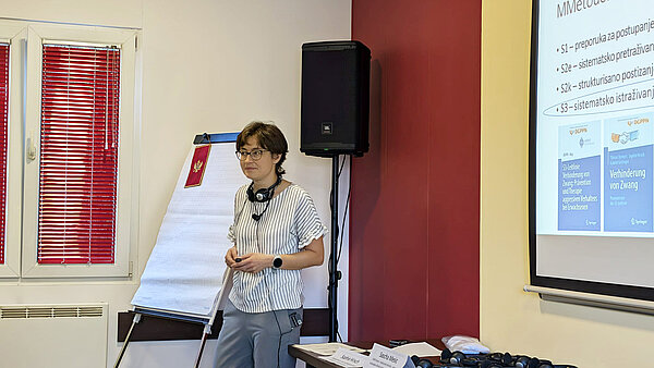 Eine junge Frau mit kurzem Haar vor einer Leinwand, auf die eien Präsentation projeziert wird. Neben ihr liegen mehrere Kopfhörer für die simultane Übersetzung.