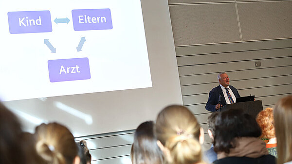 Ein Redner im dunklen Anzug steht in einer Veranstaltungshalle am Rednerpult, an der Leinwand hinter ihm ist eine PowerPoint-Präsentation zu sehen, im Vordergrund Zuhörer:innen.