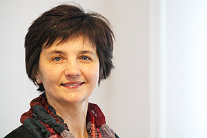 Portrait einer Frau mit dunklen halblangen Haaren, die vor einer weißen Wand steht.