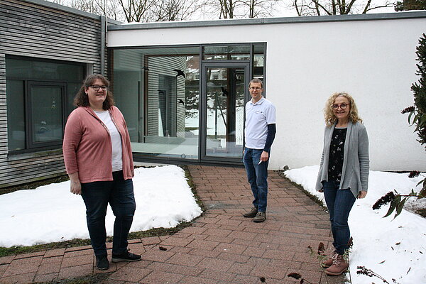 zwei Frauen im Vordergrund und ein Mann im Hintergrund stehen in Abstand zueinander draußen. Sie tragen keine Jacken, dennoch ist um sie herum Schnee zu sehen.