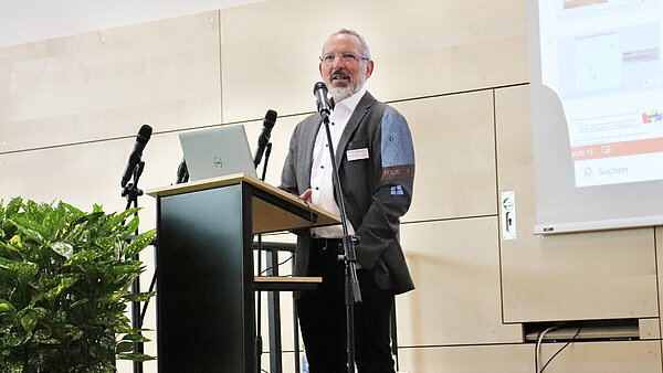 Ein Mann im Anzug mit Brille und Bart steht an einem Rednerpult und hält einen Vortrag.