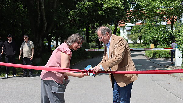 Eine Frau und ein Mann öffnen die Schleife eines roten Bandes, das über einen Weg gespannt ist. 