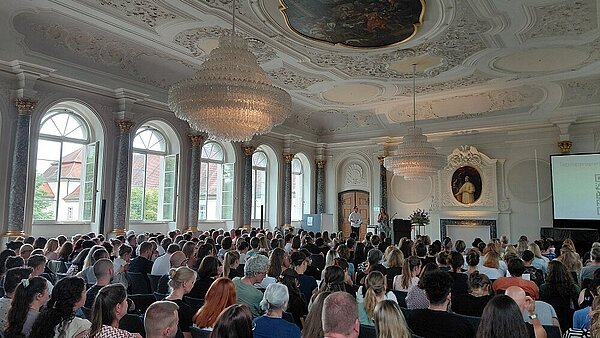 Zu sehen ist ein vollbesetzter, bestuhlter Klostersaal, fotografiert von hinten. Die Sitzenden blicken auf eine kleine Bühne nach vorne, wo zwei Personen moderieren.