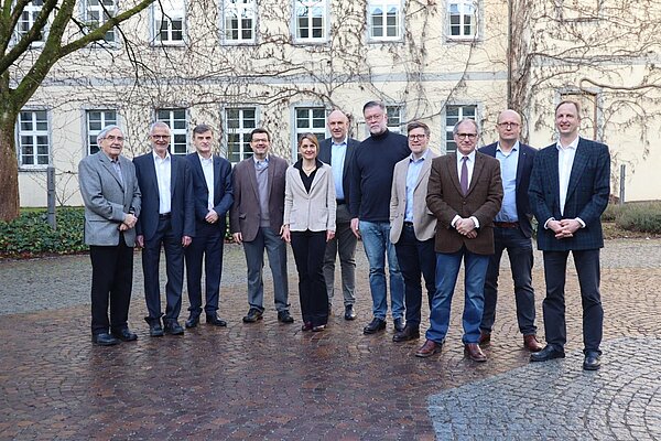 Eine Frau und zehn Männer stehen vor der Fassade eines historischen Gebäudes zum Gruppenbild zusammen und schauen freundlich in Richtung Kamera.
