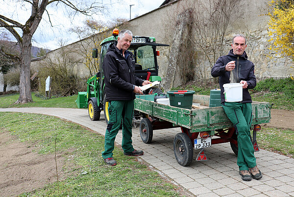 zwei Männer in Arbeitskleidung hinter einem Traktor mit Anhänger