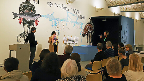 In einem Museumsraum sprechen stehend zwei Frauen und ein Mann vor sitzendem Publikum.