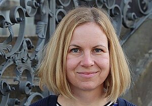 Porträtfoto einer Frau mittleren Alters mit schulterlangen blonden glatten Haaren. Sie trägt ein dunkelblaues Oberteil und lächelt freundlich in die Kamera. Im Hintergrund ist ein Treppengeländer zu sehen.