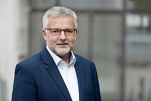 Ein Mann mit grauem Haar und Bart, einer Brille und einem dunkelblauen Sakko.