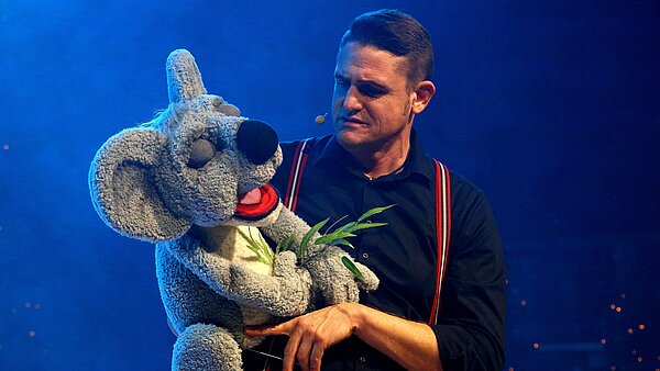 Vor einem blauen Hintergrund steht ein junger Mann mit kurzen Haaren. In seinen Händen hält er eine große Puppe in Gestalt eines Koalabären. 