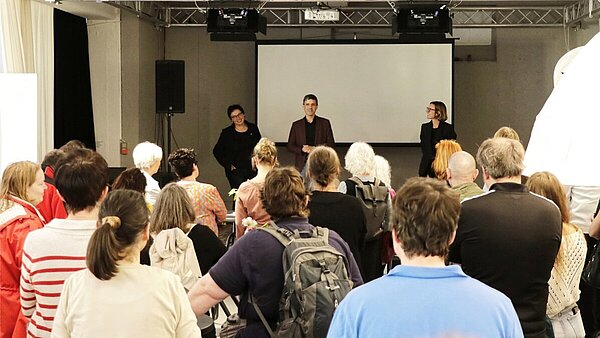 Ein Mann und zwei Frauen stehen auf der kleinen Bühne eines Veranstaltungssaals, vor ihnen stehen zahlreiche Zuhörerinnen und Zuhörer.