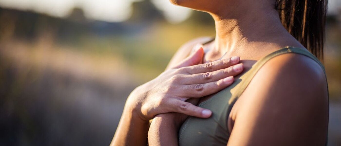 Eine junge Frau steht in der Natur und hält beide Hände flache auf die Bruste, die Lippen sind zusammengepresst.