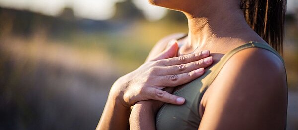 Eine junge Frau steht in der Natur und hält beide Hände flache auf die Bruste, die Lippen sind zusammengepresst.