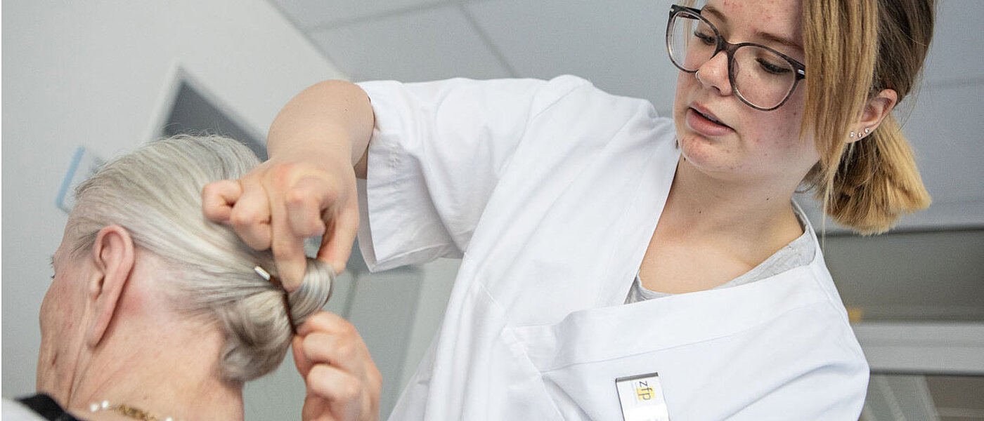 Eine junge Pflegerin mit Brille bindet gerade einer älteren Frau die Haare zusammen.