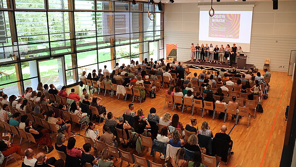 Blick von oben auf eine vollbesetzte Halle, auf der Bühne stehen mehrere referierende Personen.