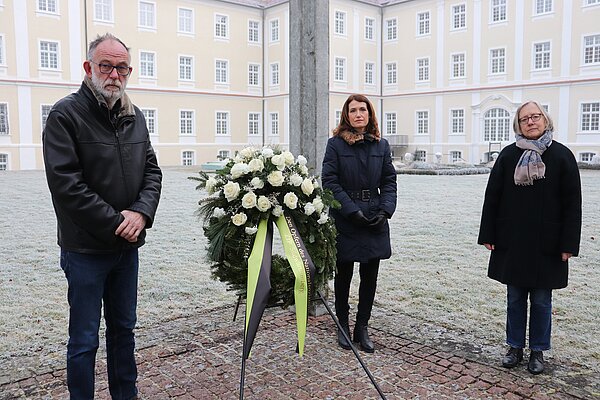 Ein Mann und zwei Frauen stehen mit ernsten Mienen an einem Trauerkranz mit dem Schleifenaufdruck "Den Opfern des Nationalsozialismus"
