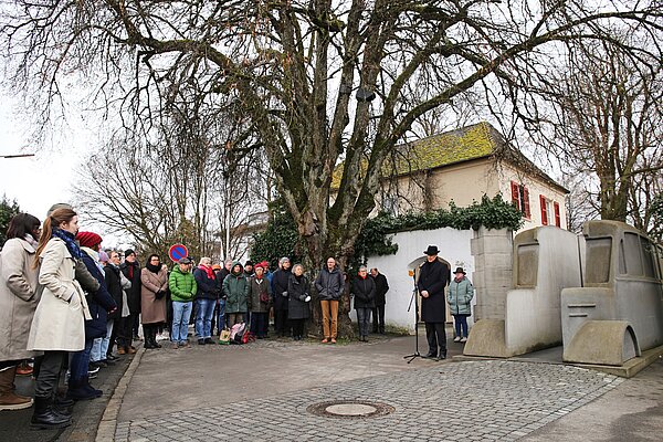 Ein schwarz gekleideter Mann steht rechts vor einem grauen Beton-Bus und hält dort eine Rede. Auf dem gepflasterten Platz vor ihm sind in einem Halbkreis links um ihn herum dutzende Menschen versammelt und hören ihm zu.